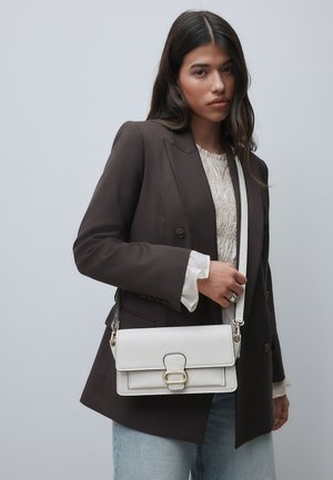 Barna blézert és fehér blúzt viselő nő arany csattal díszített fehér bőr oldaltáskát tart, sima háttér előtt áll.