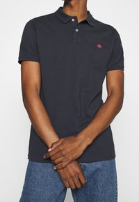 T-shirt polo bleu marine en coton, avec un col, une patte de boutonnage à deux boutons et un petit logo rouge sur la poitrine. Manches courtes.
