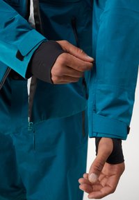 Veste imperméable couleur sarcelle avec un poignet intérieur noir. La main ajuste une fermeture éclair sur la manche, mettant en avant un design élégant et pratique.