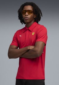 Piros galléros póló, sárga Ferrari logóval és fehér Puma logóval. A modellen borostyán színű napszemüveg van. Sima textúra.