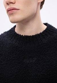 Pulover negru texturat din tricot, cu guler rotund și o suprafață moale și pufoasă. Prezintă un logo subtil la guler.