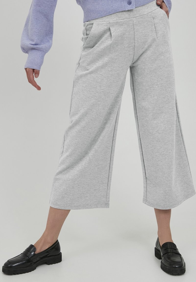 Personne portant un pantalon large gris clair raccourci, des mocassins noirs et un pull lavande, une main dans la poche.