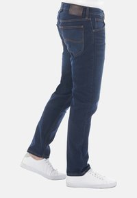 Vaqueros de mezclilla azul oscuro con corte recto, que presentan un parche de cuero con el logo en la cintura y sutiles detalles de costura. Llevados con zapatillas blancas.