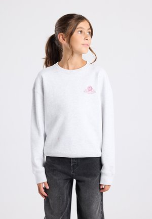 Meisje met bruin haar in een paardenstaart, gekleed in een witte sweatshirt met roze logo en zwarte jeans, kijkt opzij tegen een witte achtergrond.