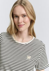 T-shirt à rayures à manches courtes en crème avec des rayures noires, présentant un col rond et un logo soleil doré sur la poitrine gauche.