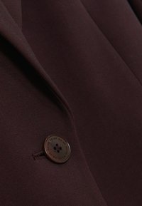 Bordinės spalvos švarkas su tekstūruota medžiaga, aštriu atvartų kraštu ir tamsiu mygtuku su reljefiniu gėlių raštu. Artimas planas pabrėžia detales.