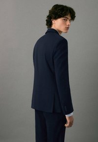 Mornarski prilagojen smoking z oblikovanim sakom, stranskimi reži, zapiranjem na en gumb in kontrastnimi svetlomodrimi manšeti. Model obrne hrbet.