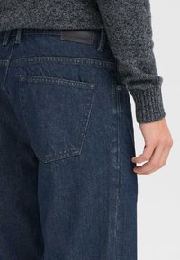 Jeans de denim oscuro con un corte recto, que cuentan con un bolsillo trasero y una etiqueta de cuero en la cinta de la cintura. La tela tiene una ligera textura.
