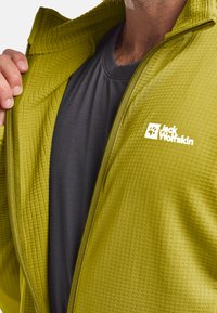 Veste zippée verte avec un motif texturé, dotée d'une couche intérieure noire. Logo blanc sur la poitrine gauche. Détail de fermeture éclair lisse.