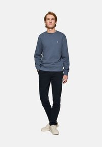 Blå crewneck sweatshirt lavet af bomuld, parret med mørke bukser og beige sneakers. Har et lille logo på brystet.