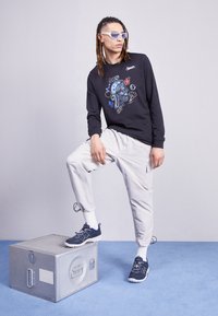 Maglietta nera a maniche lunghe con grafica colorata, pantaloni jogger grigi chiari, sneakers blu navy, modello in posa su una scatola di stoccaggio argentata contro uno sfondo semplice.