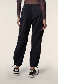 Rückansicht einer Person, die schwarze Trainingshose mit elastischem Bund und Bündchen am Knöchel sowie schwarz-weiße Sneakers vor einem einfarbigen Hintergrund trägt.