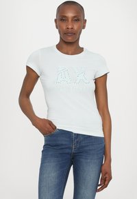 Hele sinine lühikeste varrukatega T-särk ümmarguse kaelusega, millel on studded "AX" logo. Kombineeritud tumehallide kitsaste teksadega, käed taskutes.