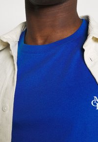 Blaues Rundhals-T-Shirt, figurbetont, getragen unter einem hellbeigen Kordhemd mit Knopfleiste, das ein weißes Logodetail auf der Brust zeigt.