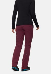 Pantaloni da esterno bordeaux con design affusolato, caratterizzati da dettagli neri e logo, abbinati a un top lungo a maniche nere con texture e scarpe multicolori.