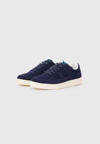 Sneakers din suede bleumarin cu talpă din cauciuc alb, șireturi plate și accente verzi pe limbă. Au detalii de cusătură și o suprafață texturată.