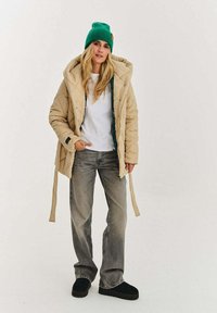 Béžová prošívaná péřová bunda s kapucí a zelenou podšívkou. Kombinována s volnými šedými džínami a zelenou pletenou čepicí. Černé slip-on boty.