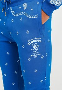 Niebieskie spodnie dresowe z białymi wzorami paisley, teksturowaną tkaniną, ściągaczem z troczkiem w talii oraz logo na udzie.