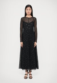 Čierne šaty s dlhými priehľadnými rukávmi, s bodkovaným vzorom, dĺžkou po zem a zaobleným výstrihom. Kombinované s čiernymi sandálmi na remienkoch.