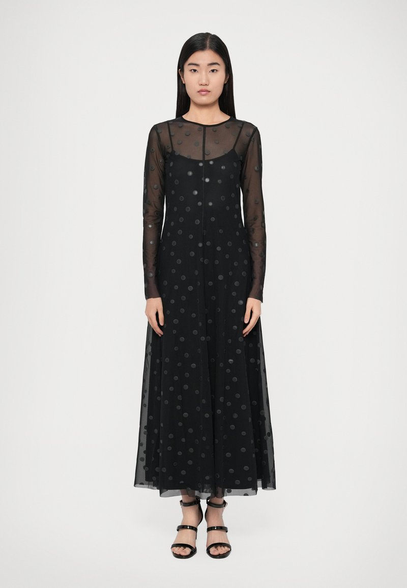 Čierne šaty s dlhými priehľadnými rukávmi, s bodkovaným vzorom, dĺžkou po zem a zaobleným výstrihom. Kombinované s čiernymi sandálmi na remienkoch.