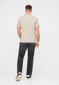 Beiges T-Shirt mit kurzen Ärmeln und Rundhalsausschnitt, kombiniert mit dunkelgrauen Jeans und schwarzen Sneakers; das Outfit zeichnet sich durch einen lässigen Schnitt und klare Linien aus.