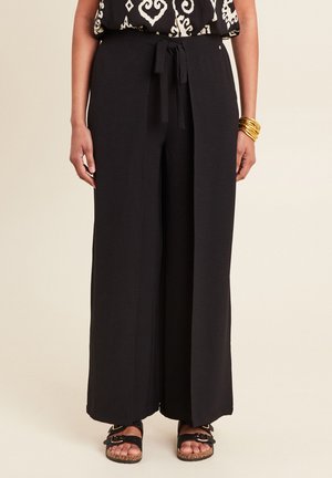 Pantalons larges noirs en tissu texturé, avec une taille à nouer et des poches latérales, associés à des sandales noires et des bracelets en or.