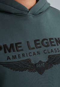 Zelena dukserica s crnim vezom "PME Legend American Classic" i logom s krilatim štitom na prsima, koju nosi osoba.