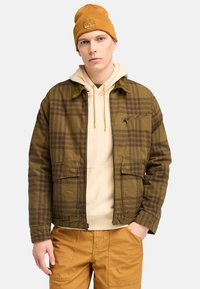 Brun ternet jakke med en lynlås lomme, over en beige hættetrøje. Modellen bærer en sennepsgul beanie. Outfitet inkluderer brune cargo bukser.