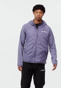 Young man wearing a purple quilted zip-up jacket with hood and black pants, standing against a plain white background.