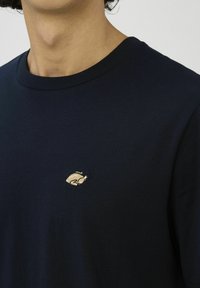 Homme portant un t-shirt bleu marine foncé avec un petit patch brodé représentant un poulet rôti sur le côté gauche de la poitrine.
