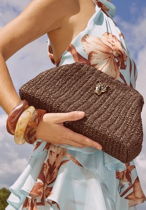 Žena v kvetovanom šatke na uväzovanie okolo krku drží pletenú hnedú listovú kabelku so zapínaním v tvare leopardej hlavy, nosí hrubé jantárové náramky na pozadí modrej oblohy.