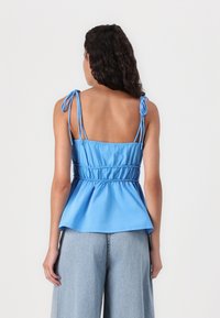 Blue cotton peplum top with adjustable tie straps, smocked waist, and a soft texture. Paired with light blue wide-leg jeans.