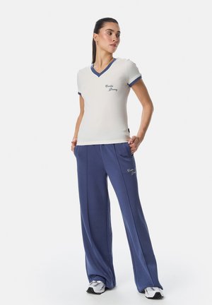 Žena stojí s rukama v kapsách, nosí bílý tričko s výstřihem do V a volné modré kalhoty, obojí s nápisem "Cardio Bunny", a bílé tenisky.