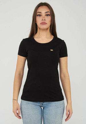Donna con lunghi capelli castani che indossa una semplice maglietta nera a maniche corte e jeans azzurri chiari, in piedi davanti a uno sfondo bianco uniforme.