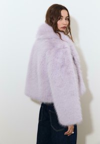 Lilla faux fur cropped jakke med lange ærmer og høj krave, der har en blød, fluffy tekstur. Båret over mørke jeans.