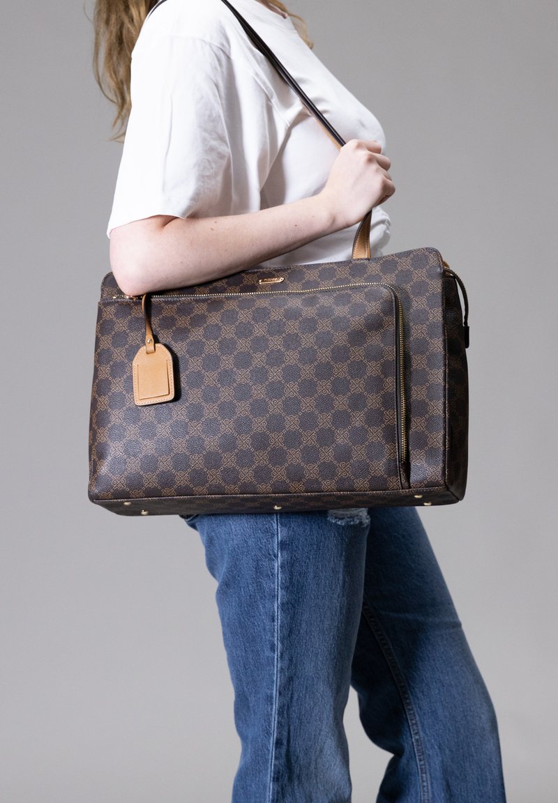 Bruine tote bag met een gestructureerd ontwerp, leren accenten en een afneembaar bagagelabel, gedragen op een schouder.