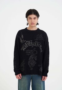 Maglione nero a coste con grafiche in un colore più chiaro. Vestibilità oversize con maniche lunghe e scollatura a girocollo, abbinato a jeans in denim sbiadito.