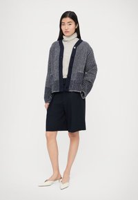 Pletený kardigán s vertikálnymi pruhmi v námorníckej modrej a sivej farbe, vybavený zapínaním na gombíky, doplnený priliehavým rolákom a nohavicami na mieru.