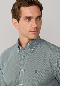 Camisa de cuadros verdes y blancos con botones, que presenta un cuello puntiagudo, una tapeta frontal con botones y un pequeño logotipo bordado en el pecho.