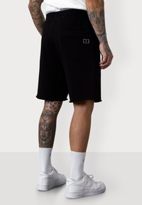 Shorts en coton noirs avec une taille élastique, comprenant une poche arrière et un logo coloré. Des baskets blanches et des chaussettes complètent la tenue.