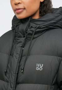 Pessoa a usar uma jaqueta acolchoada preta da HUGO com colarinho alto e cordões, lábios visíveis, parte do rosto mostrado.