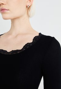 Top preto canelado com decote de rendas em forma de onda, com corte justo e mangas longas. Vista em close que enfatiza a textura e os detalhes.