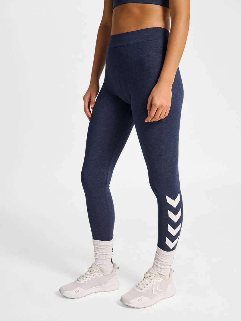 Frau trägt marineblaue Leggings mit weißen Chevron-Streifen an den Waden, weiße Socken und weiße Sportschuhe, steht auf einer weißen Oberfläche.
