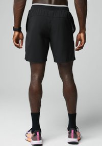 Schwarze Sportshorts mit einem grauen Bund, ausgestattet mit seitlichen Taschen. Das Modell trägt bunte Turnschuhe und schwarze Socken.