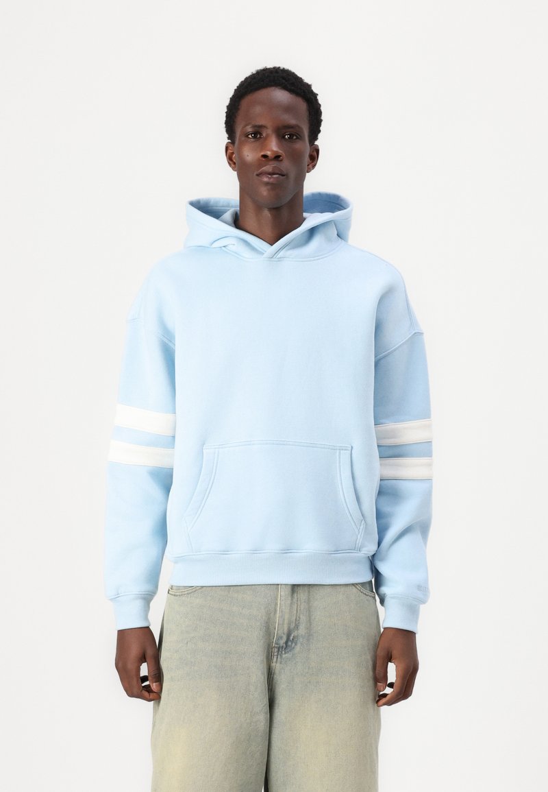 Světle modrá mikina s kapucí, přední kapsou, žebrovanými manžetami a třemi bílými pruhy na levém rukávu. Měkká látka, volný střih, klasický design.