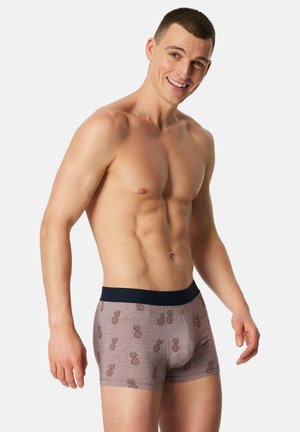 Brune boxershorts med små ananasmønstre, en marineblå linning, lavet af et blødt stof. Modellen står med et neutralt udtryk.
