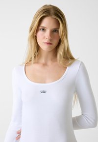 Biała, prążkowana bluzka z długim rękawem, z okrągłym dekoltem i małym czarnym logo "GUESS JEANS" na przodzie. Gładka faktura.