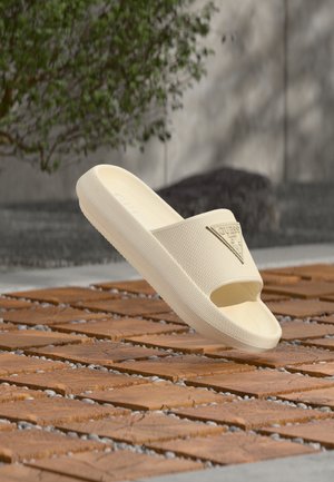 Sandale slip-on bej cu talpă texturată și logo GUESS pe baretă, plutind deasupra unei suprafețe exterioare din plăci de lemn și pietre.