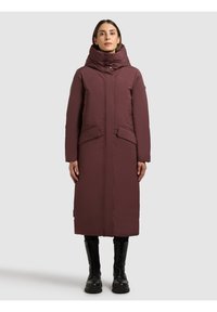 Hosszú, bordó pufi kabát magas gallérral, nagy első zsebekkel és egyenes szabással. Fekete bakancsokkal párosítva a kontraszt érdekében.