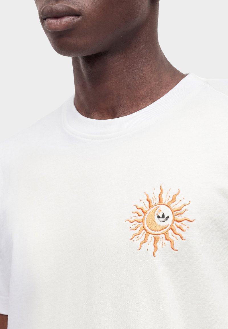 Primo piano di una persona che indossa una T-shirt bianca con un logo arancione di sole e luna crescente sul lato sinistro del petto.